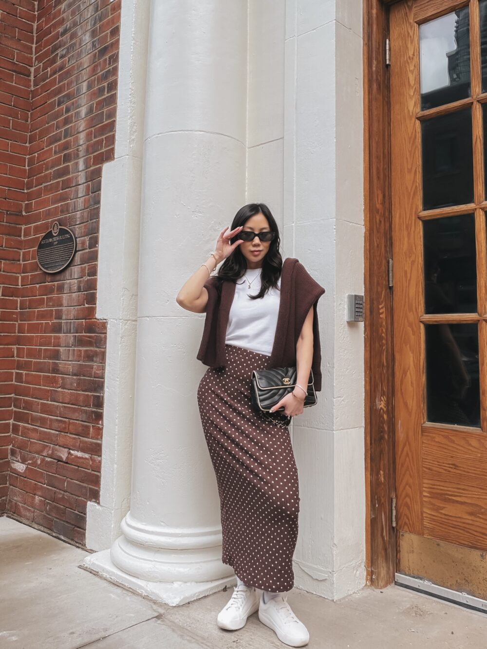 yesmissy wearing a chocolate brown polka dot maxi skirt, white tee, caramel sweater on shoulders and white sneakers